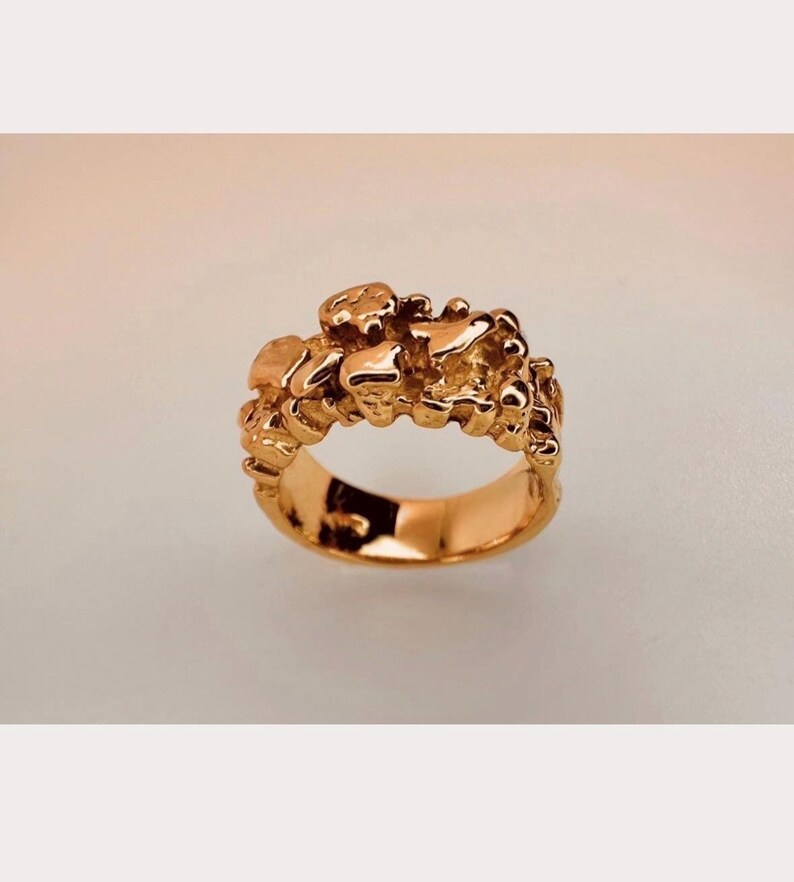 May include: A gold ring featuring a band with a highly textured, irregular surface composed of multiple raised, rounded, and molten-looking metallic shapes. The metal has a polished, reflective finish that catches the light across its uneven, organic form.