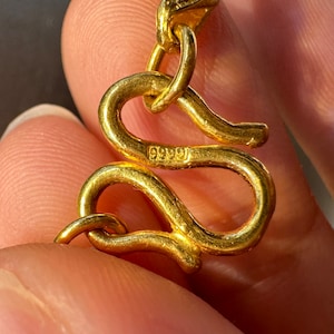 May include: Close-up of a gold-colored jewelry clasp. The clasp is in the shape of a stylized snake. The clasp has the number "9999" stamped on it. The jewelry is likely a necklace or bracelet.