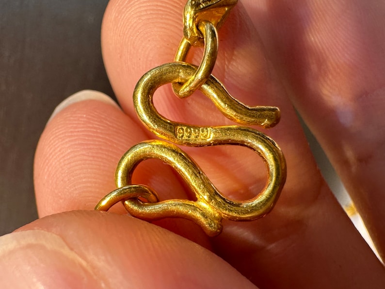 May include: Close-up of a gold-colored jewelry clasp. The clasp is in the shape of a stylized snake. The clasp has the number "9999" stamped on it. The jewelry is likely a necklace or bracelet.