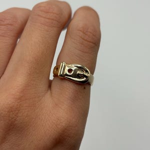 A gold-colored ring in the shape of a belt buckle. The ring is worn on a finger and has a small, round hole in the buckle.