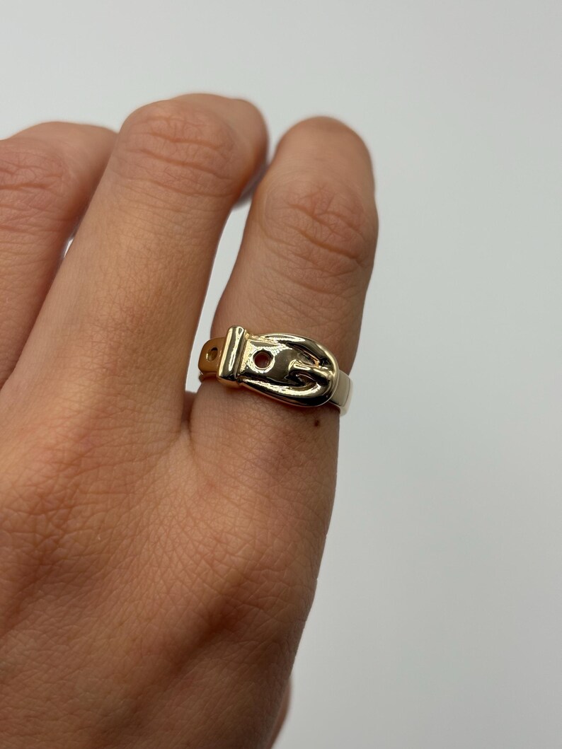 A gold-colored ring in the shape of a belt buckle. The ring is worn on a finger and has a small, round hole in the buckle.