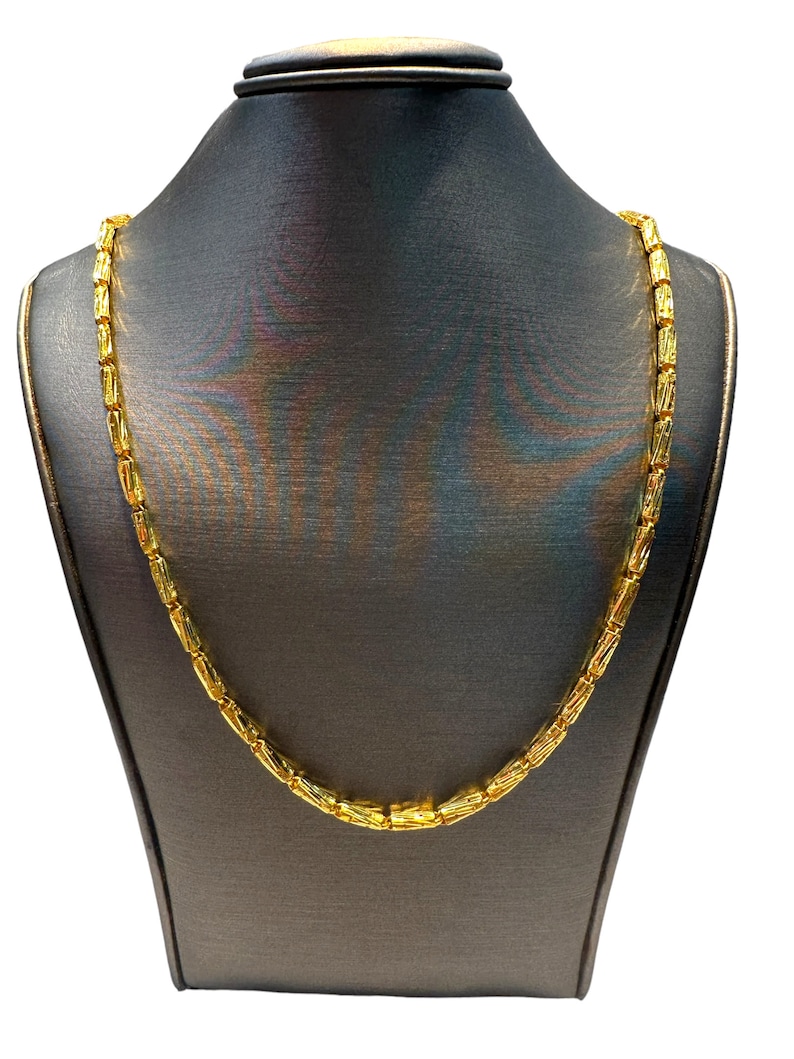 May include: A gold-colored necklace with a rectangular link design is displayed on a dark gray jewelry bust. The necklace is made of small, rectangular links that are connected to form a chain. The jewelry bust is a neutral backdrop.