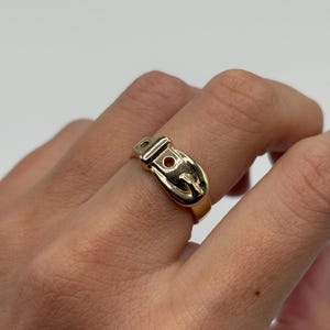 A gold-colored ring in the shape of a belt buckle. The ring is worn on a finger and has a shiny, polished finish. The buckle design includes a small, circular detail.
