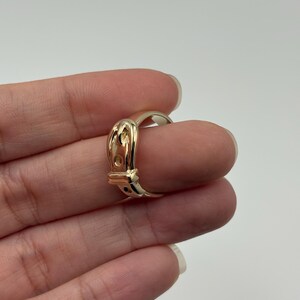 A gold-colored ring with a buckle design. The ring features a detailed buckle shape with a small, rectangular opening and a curved band. The ring is shown on a finger against a plain background.
