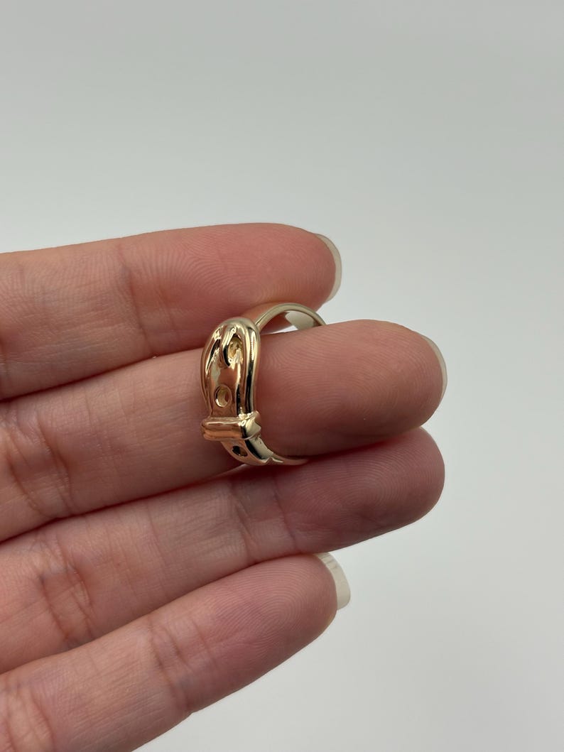 A gold-colored ring with a buckle design. The ring features a detailed buckle shape with a small, rectangular opening and a curved band. The ring is shown on a finger against a plain background.