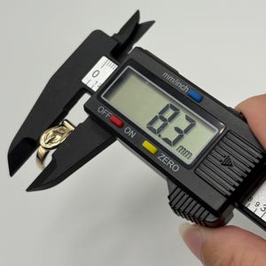 A gold-colored ring is measured with a black digital caliper. The caliper displays a measurement of 8.3 mm.
