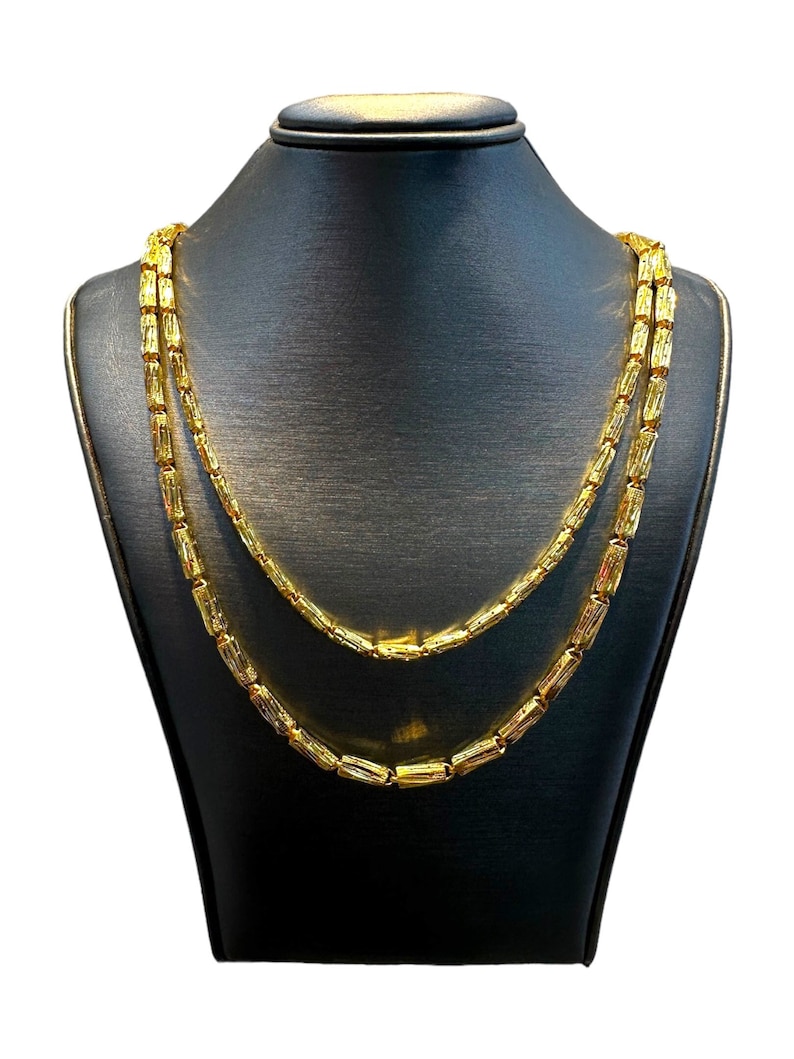 May include: A gold-colored necklace with a double-strand design. The necklace is displayed on a black mannequin. The necklace features rectangular, tube-shaped beads. The jewelry is presented against a white background.