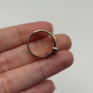 A gold-colored ring with a decorative belt design. The ring is shown on a hand against a white background.