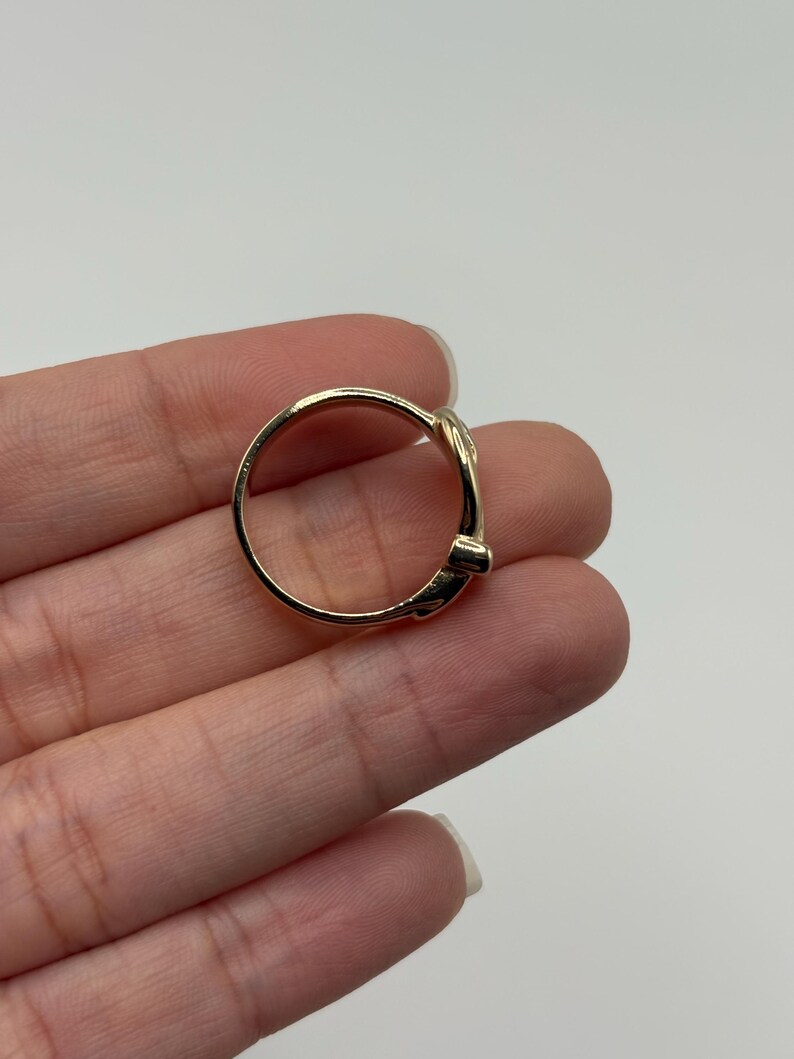 A gold-colored ring with a decorative belt design. The ring is shown on a hand against a white background.