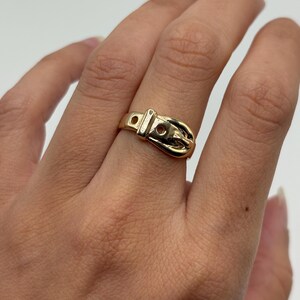 A gold-colored ring in the shape of a belt buckle. The ring has a detailed design with a buckle and holes, and is worn on a finger. The ring is made of 14K gold.