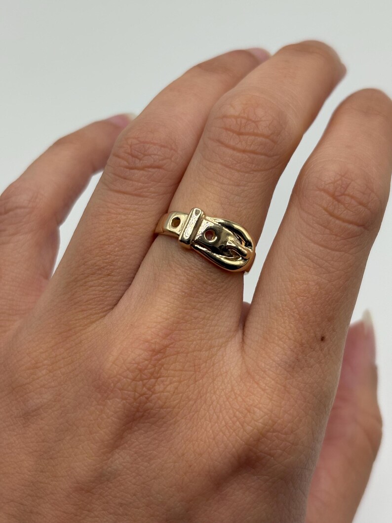 A gold-colored ring in the shape of a belt buckle. The ring has a detailed design with a buckle and holes, and is worn on a finger. The ring is made of 14K gold.