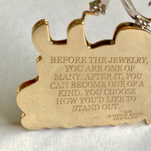 May include: Gold-colored metal pendant with an inscription that reads "Before the jewelry, you are one of many. After it, you can become one of a kind. You choose how you'd like to stand out."  The pendant also has the text "EM A GOLD MIND JEWELRY" on it.