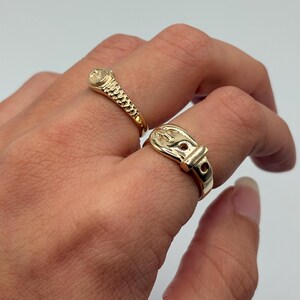 Two gold-colored rings are displayed. One ring features a textured band and a small, round stone. The other ring is designed to resemble a belt buckle. The rings are on a hand against a white background.