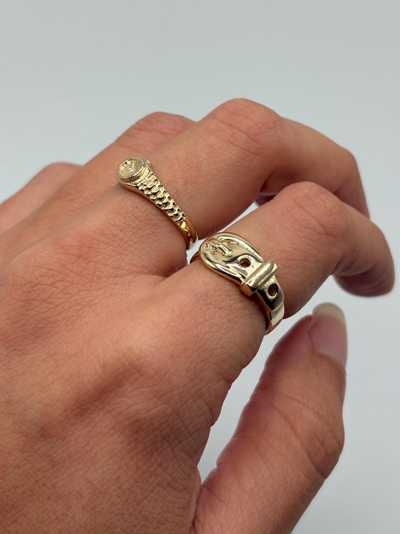 Two gold-colored rings are displayed. One ring features a textured band and a small, round stone. The other ring is designed to resemble a belt buckle. The rings are on a hand against a white background.