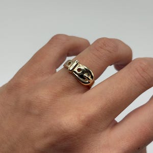 A gold-colored ring in the shape of a belt buckle. The ring is worn on a finger and has a shiny, polished finish. The buckle design includes a rectangular frame and a rounded buckle tongue.