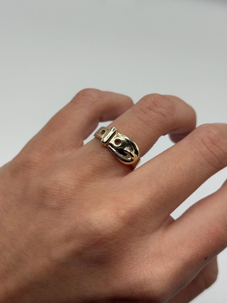 A gold-colored ring in the shape of a belt buckle. The ring is worn on a finger and has a shiny, polished finish. The buckle design includes a rectangular frame and a rounded buckle tongue.