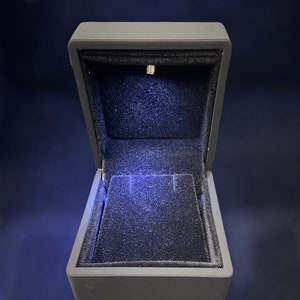 May include: A black velvet ring box with a built-in LED light. The box is open and the light is illuminating the interior.