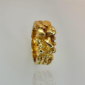 May include: A gold ring with a textured, organic surface featuring irregular, raised molten-like shapes. The band has a high-shine, reflective finish that catches the light, creating a series of uneven, rounded protrusions across the entire outer circumference.