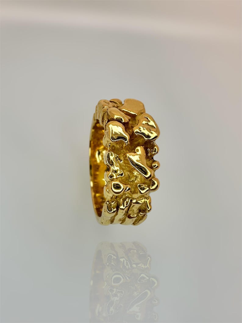 May include: A gold ring with a textured, organic surface featuring irregular, raised molten-like shapes. The band has a high-shine, reflective finish that catches the light, creating a series of uneven, rounded protrusions across the entire outer circumference.