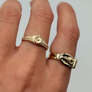 Two gold-colored rings are displayed on a hand. One ring is designed to look like a watch, and the other ring is designed to look like a belt buckle. The rings are set against a neutral background.