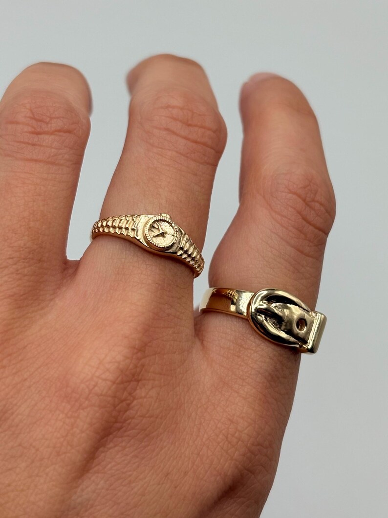 Two gold-colored rings are displayed on a hand. One ring is designed to look like a watch, and the other ring is designed to look like a belt buckle. The rings are set against a neutral background.
