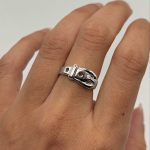 May include: A polished silver-toned ring designed to resemble a miniature belt, featuring a central buckle element, a fixed loop, and two small circular holes along the band, creating a continuous metallic loop around the finger.