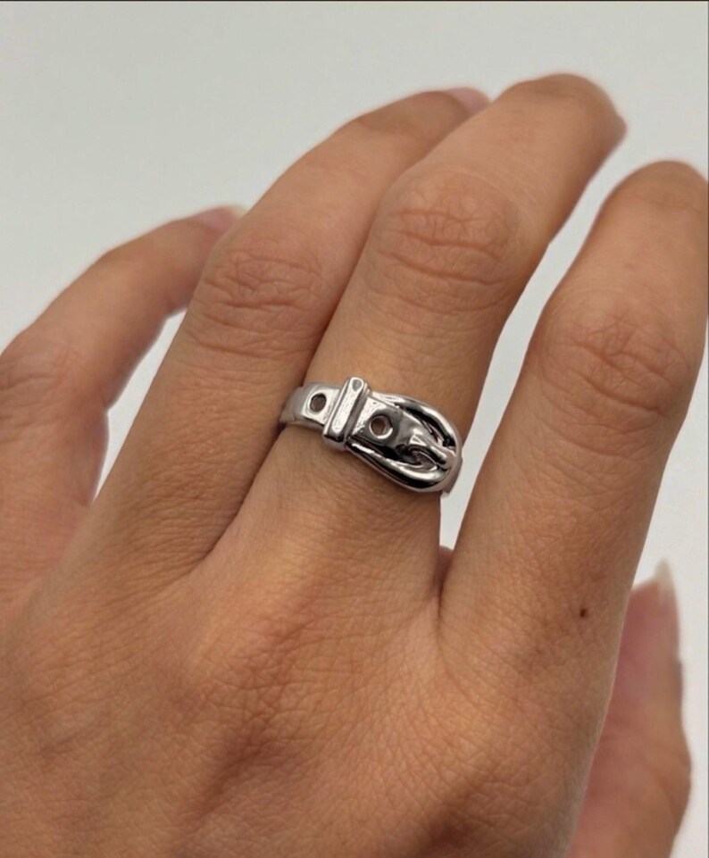 May include: A polished silver-toned ring designed to resemble a miniature belt, featuring a central buckle element, a fixed loop, and two small circular holes along the band, creating a continuous metallic loop around the finger.