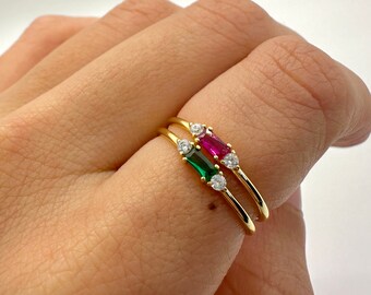 May include: Two gold rings with small, clear stones and a larger, rectangular stone. One ring has a green stone and the other has a red stone.