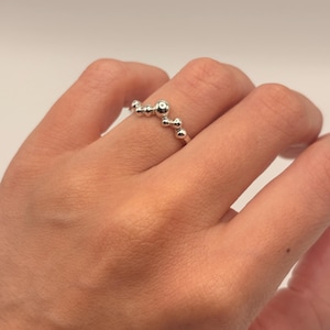 May include: A silver-toned ring featuring a series of small, polished spherical beads arranged in a curved, arching pattern along the top of the band. The metal has a reflective, smooth finish, and the beads vary slightly in size across the arc.