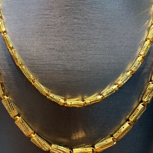 May include: Two gold necklaces with a repeating cylindrical design. The necklaces are displayed on a dark gray surface. The gold color is bright and shiny, reflecting light. The necklaces are likely made of metal.