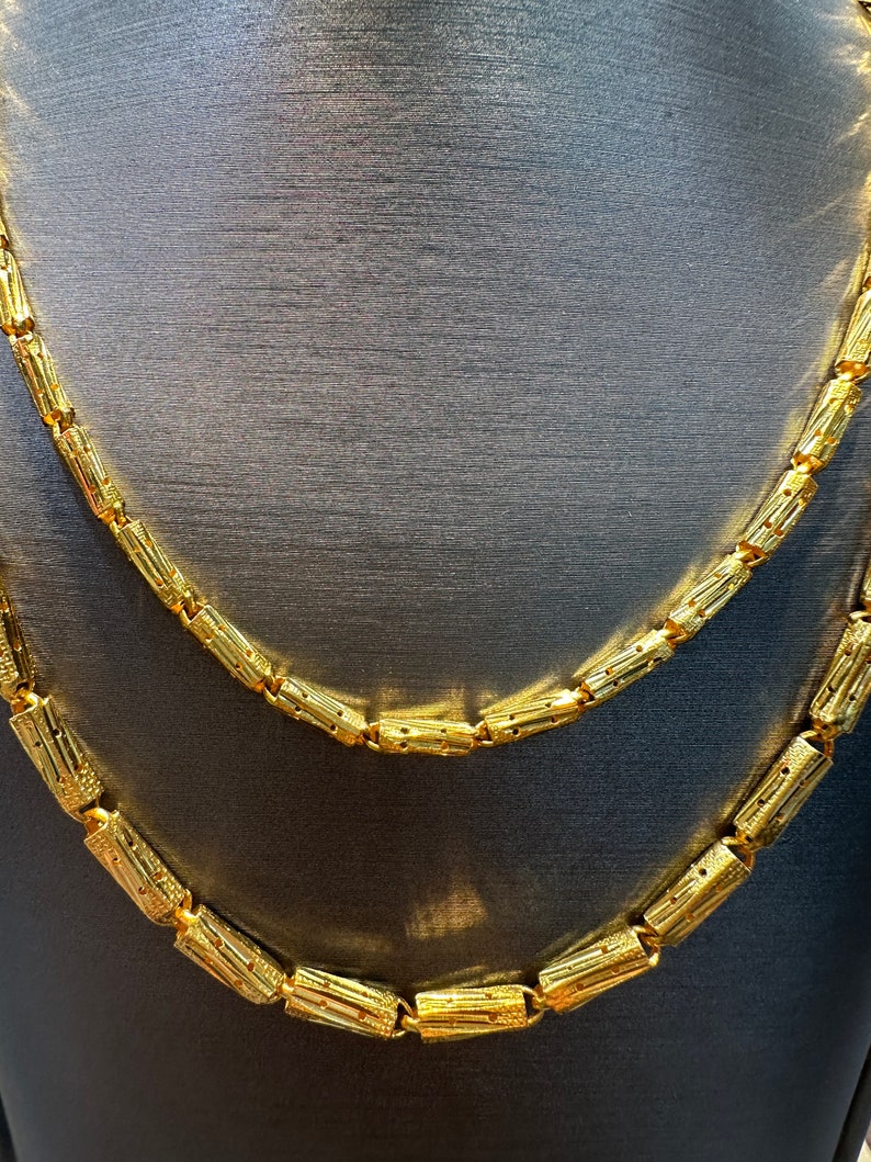 May include: Two gold necklaces with a repeating cylindrical design. The necklaces are displayed on a dark gray surface. The gold color is bright and shiny, reflecting light. The necklaces are likely made of metal.