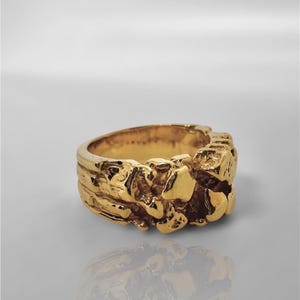 May include: A gold ring with a thick, band-style design featuring a highly textured, irregular, and organic surface that resembles molten metal or raw nuggets. The finish is polished and reflective, creating highlights across the uneven surface.