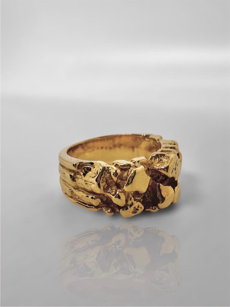 May include: A gold ring with a thick, band-style design featuring a highly textured, irregular, and organic surface that resembles molten metal or raw nuggets. The finish is polished and reflective, creating highlights across the uneven surface.