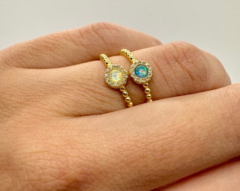 May include: Two gold rings with opal gemstones set in a halo of small clear stones. The rings are on a finger.