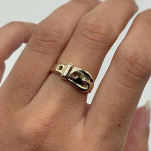 A gold-colored ring in the shape of a belt buckle. The ring is worn on a finger and has a detailed design, including a buckle and holes.
