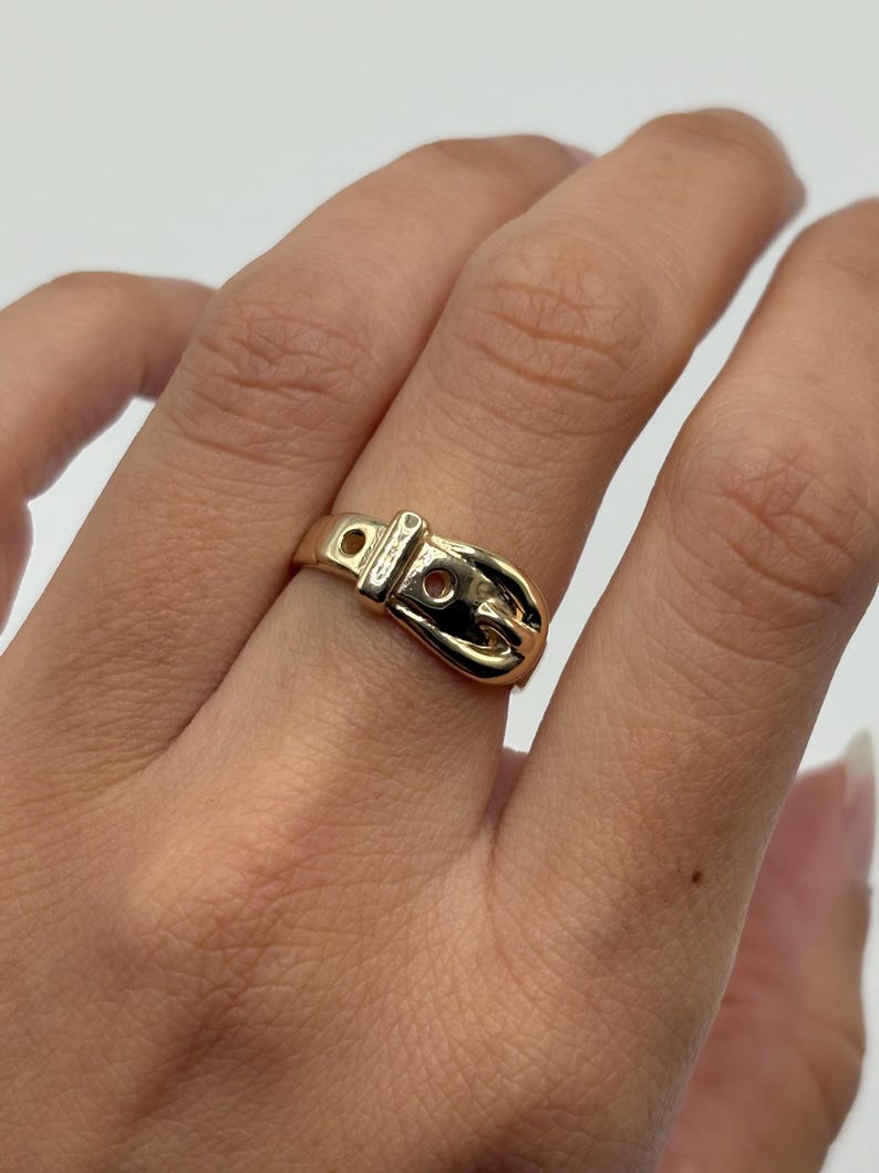 A gold-colored ring in the shape of a belt buckle. The ring is worn on a finger and has a detailed design, including a buckle and holes.