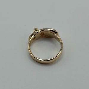 A gold-colored ring with a rounded band and a decorative top. The top of the ring features a small, raised design. The ring is set against a plain, light background.