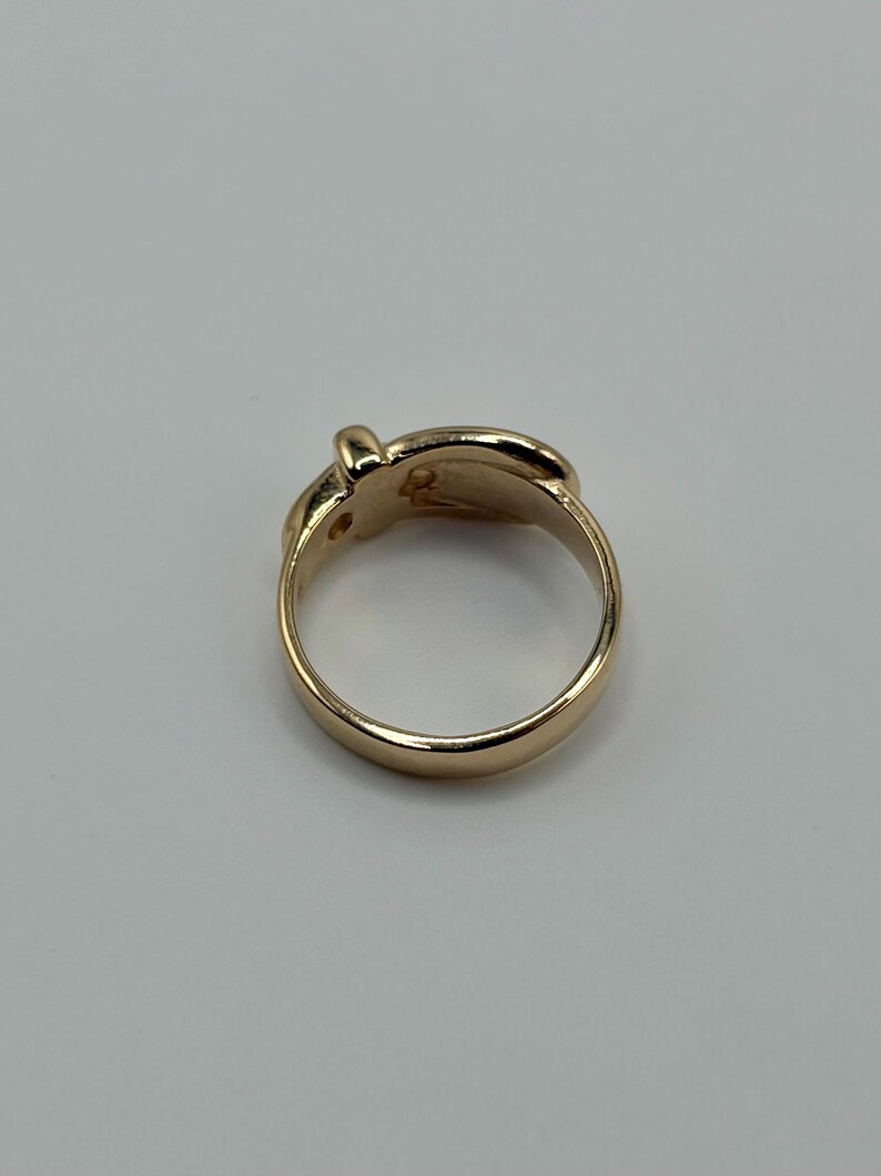 A gold-colored ring with a rounded band and a decorative top. The top of the ring features a small, raised design. The ring is set against a plain, light background.