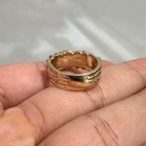 May include: A gold-toned ring with a polished, smooth band and a textured, uneven top edge. The metal surface reflects light, showing a warm, metallic yellow hue throughout the circular structure of the jewellery piece.
