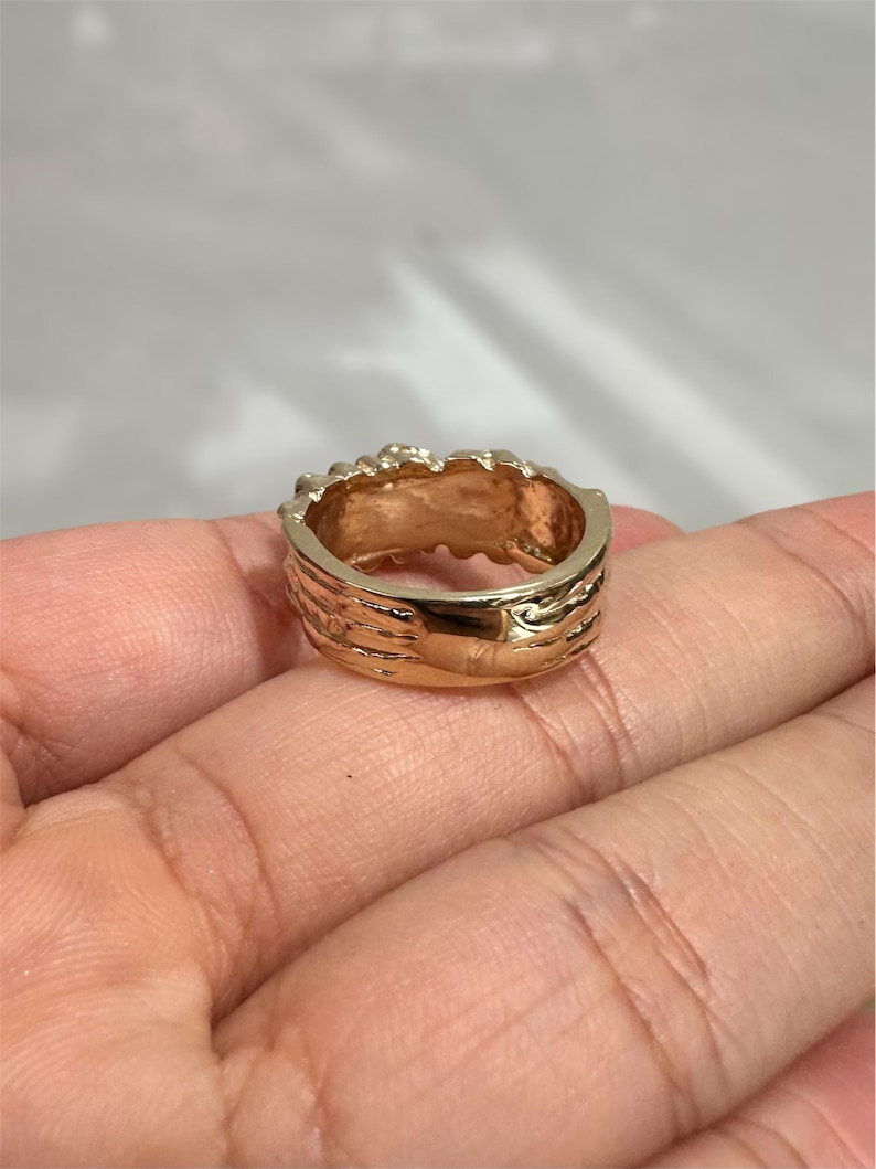 May include: A gold-toned ring with a polished, smooth band and a textured, uneven top edge. The metal surface reflects light, showing a warm, metallic yellow hue throughout the circular structure of the jewellery piece.