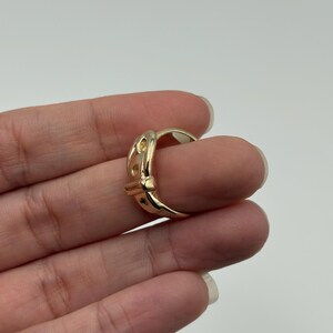 A gold-colored ring with a buckle design. The ring features a series of small, evenly spaced cutouts along the buckle's surface. The ring is worn on a finger, showcasing its polished finish and intricate details.