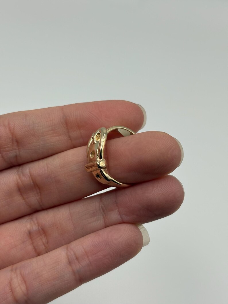 A gold-colored ring with a buckle design. The ring features a series of small, evenly spaced cutouts along the buckle's surface. The ring is worn on a finger, showcasing its polished finish and intricate details.