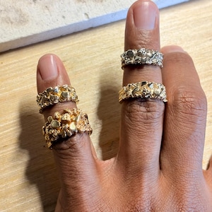 May include: Four rings with a textured, nugget-like surface finish. Two rings are gold-toned and worn on the index finger, while one gold-toned ring and one silver-toned ring are worn on the middle finger. Each ring features an irregular, organic shape.