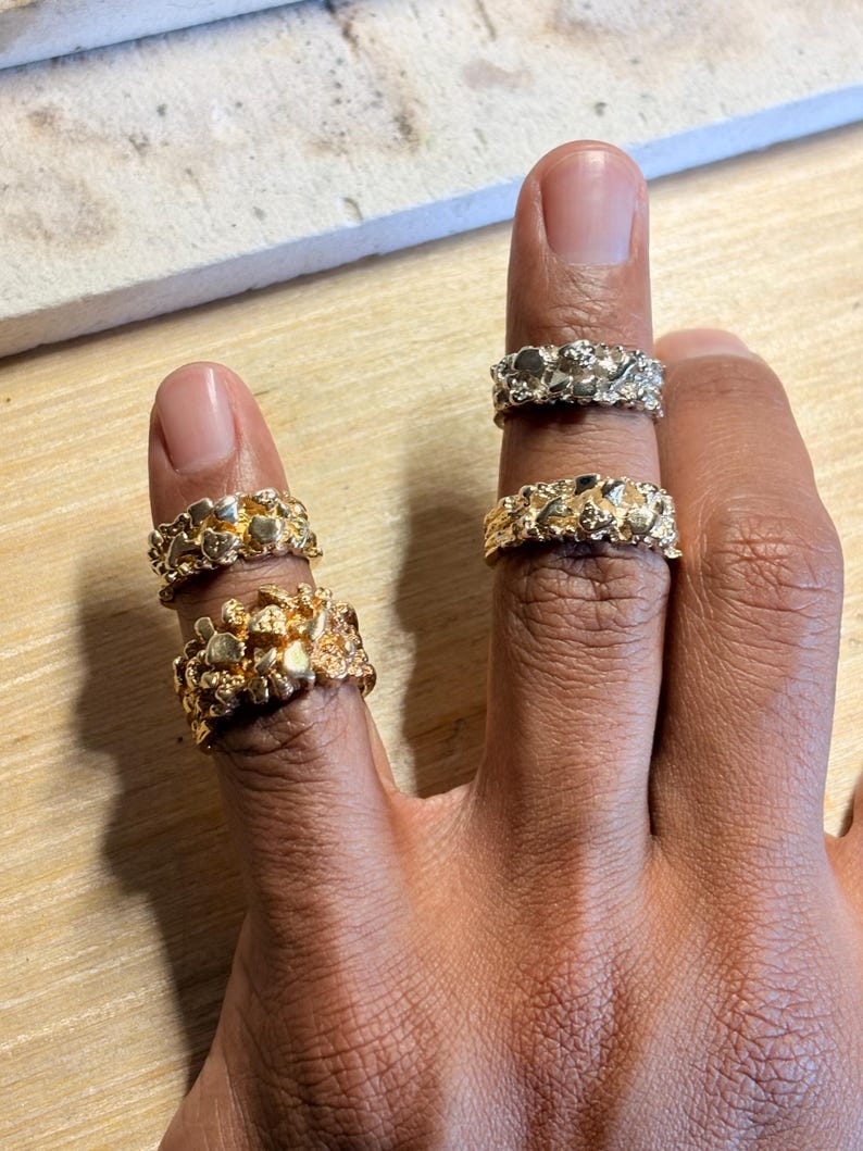 May include: Four rings with a textured, nugget-like surface finish. Two rings are gold-toned and worn on the index finger, while one gold-toned ring and one silver-toned ring are worn on the middle finger. Each ring features an irregular, organic shape.