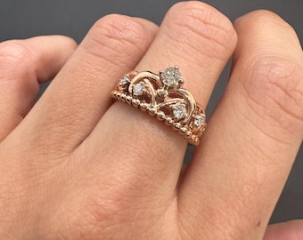May include: A rose gold crown ring with a central diamond and smaller diamonds. The ring has a delicate, ornate design with a beaded band and swirling details, creating a regal and elegant appearance.