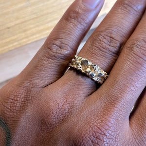 May include: A gold ring with a textured, uneven surface that resembles a natural nugget. The band features a series of raised, irregular metallic bumps and crevices that create a rugged, organic appearance across the entire circumference of the ring.