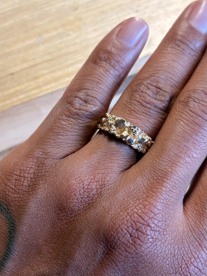 May include: A gold ring with a textured, uneven surface that resembles a natural nugget. The band features a series of raised, irregular metallic bumps and crevices that create a rugged, organic appearance across the entire circumference of the ring.