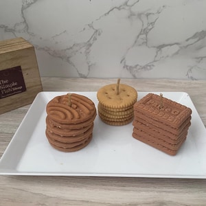 May include: A white rectangular plate holds three stacks of candles shaped like cookies and crackers. The candles are brown and tan, with wicks on top. The Simple Flair Shop logo is visible in the background.