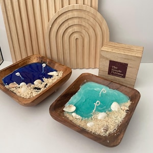 May include: Two wooden bowls with beach-themed candles. One candle is blue, the other is turquoise, both with seashells and sand. A wooden box with text and wooden decorative pieces are in the background.