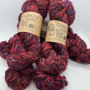 Op de afbeelding: Twee strengen Closkelt Yarns, een handgesponnen garen, in een rijke, dieprode kleur met spikkels van blauw, oranje en zwart. Het garen is gelabeld met de tekst "CLOSKELT YARNS" en "HANDSPUN IN CO. DOWN".